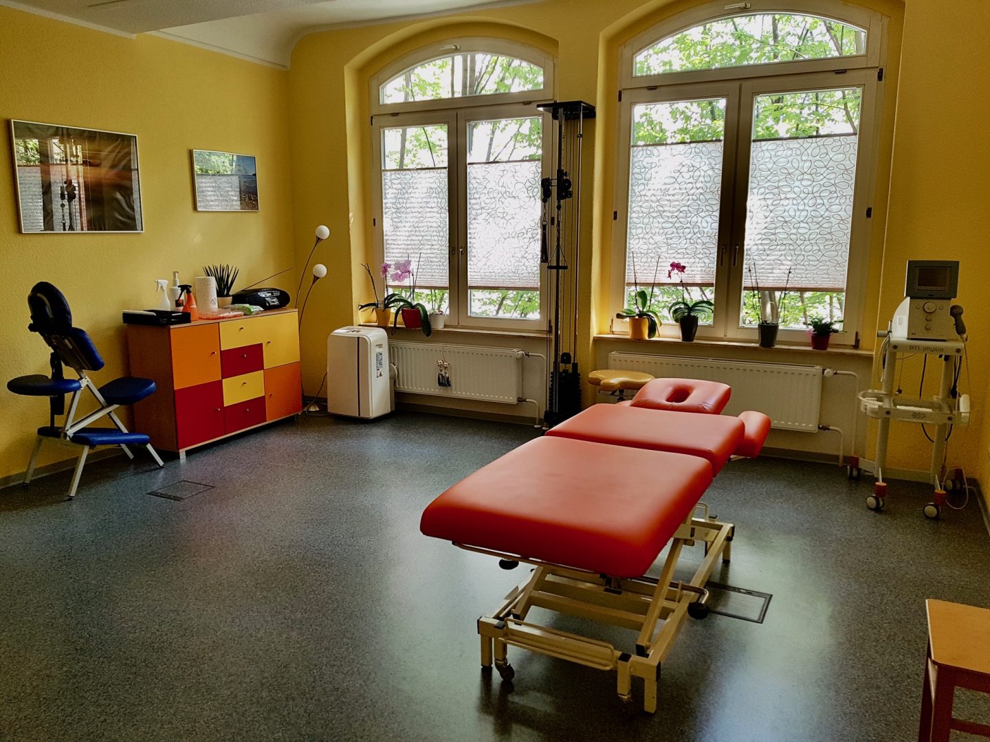 Heller Behandlungsraum der Praxis mit roter Therapieliege und großen Fenstern.